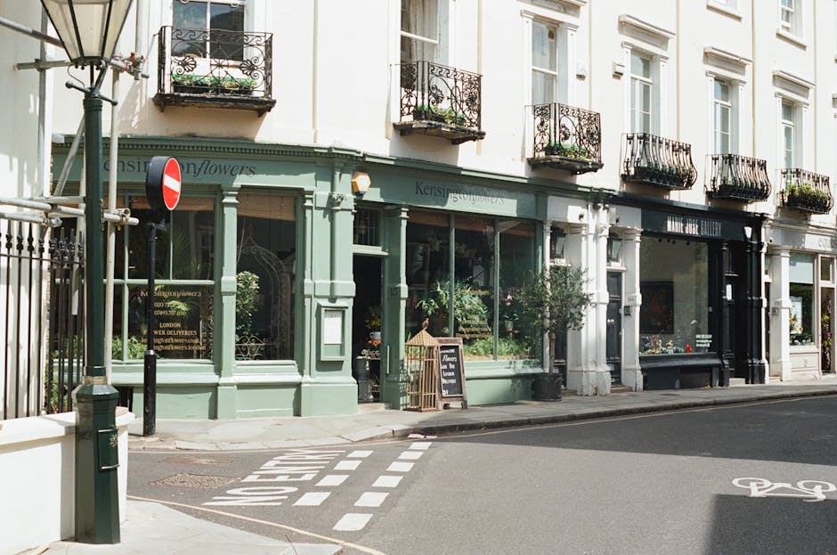 A charming flower shop with a pale green facade and large glass windows displaying vibrant floral arrangements, situated on a corner of a quiet street near Belsize Park station. The shop has decorativ