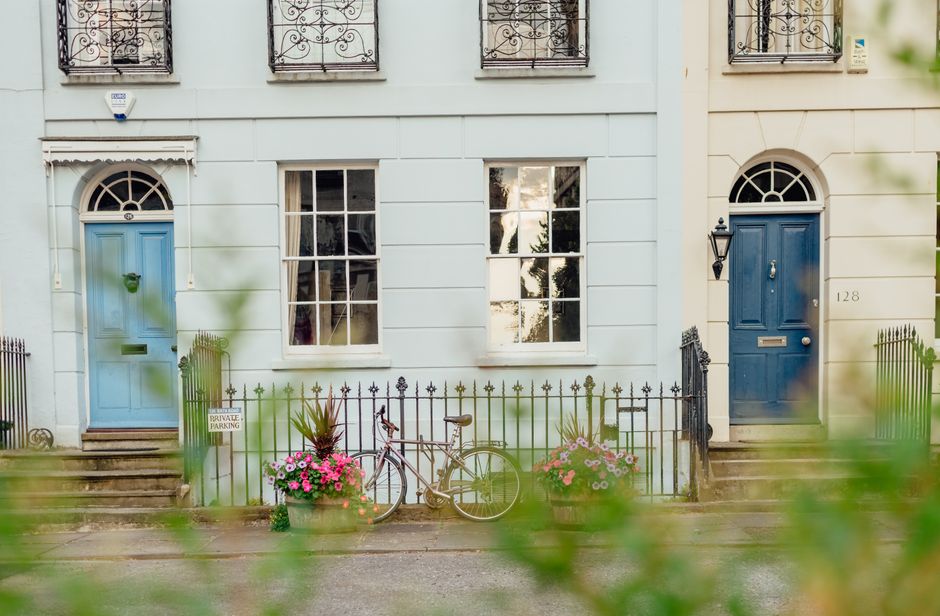 A charming residential street scene featuring two adjoining houses with pastel blue and cream facades, each with a distinct arched and rectangular window beside a vibrant blue front door. The doors ar