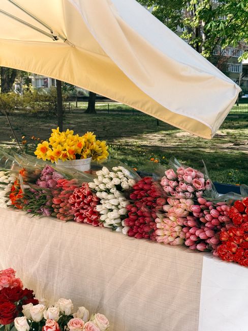 An outdoor floral market stall featuring an assortment of fresh flower bouquets arranged on a beige tablecloth under a large cream-colored umbrella. The bouquets include pink tulips, white tulips, red