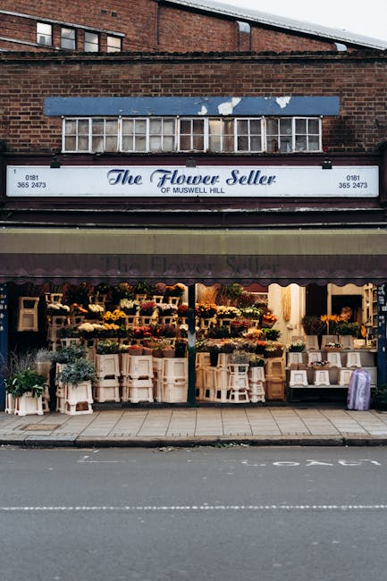 The exterior of The Flower Seller of Muswell Hill, showcasing a storefront full of vibrant floral arrangements displayed on white wooden tables and shelves. The arrangement includes a variety of fresh