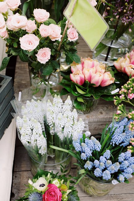 A variety of fresh floral arrangements displayed on a wooden surface, including a bouquet of soft pink roses in a clear glass vase, a cluster of white lily of the valley stems in plastic sleeves, a bu