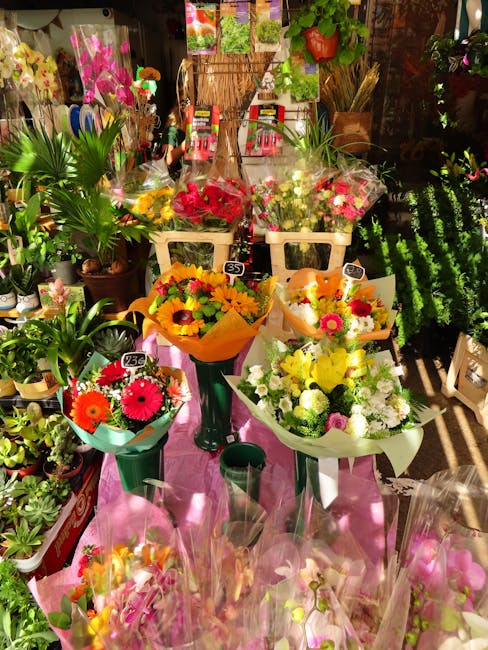 A display of vibrant, fresh flower bouquets arranged on a pink table in a floral shop. The arrangement includes a bouquet of yellow and orange flowers wrapped in orange paper, a mixed bouquet of white